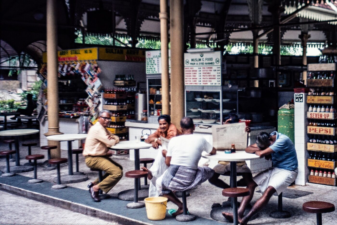 Colour photograph of a covered hawker centre with cooked-food stalls, drinks crates and customers seated at round tables.