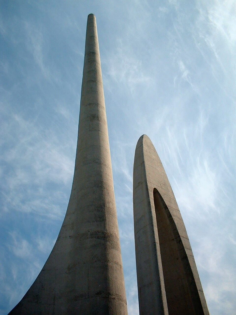 A low-angle view of two tapering concrete columns of the monument, one solid and one pierced by a tall arch, against a wispy blue sky.