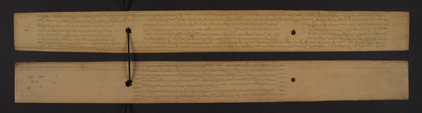 Two long narrow strips of dried palm leaf laid on a dark ground, threaded on a cord through a hole near the centre, each covered with close rows of small rounded Sinhala characters.