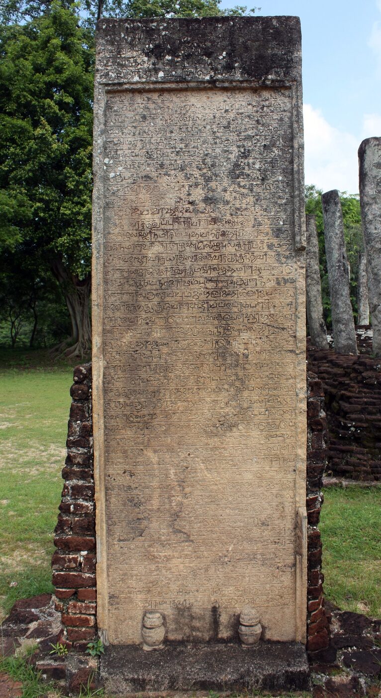 A tall weathered stone slab set upright on a brick footing in a grassy clearing, its face covered with many close-packed lines of rounded Tamil letters, with trees and further stone pillars behind.