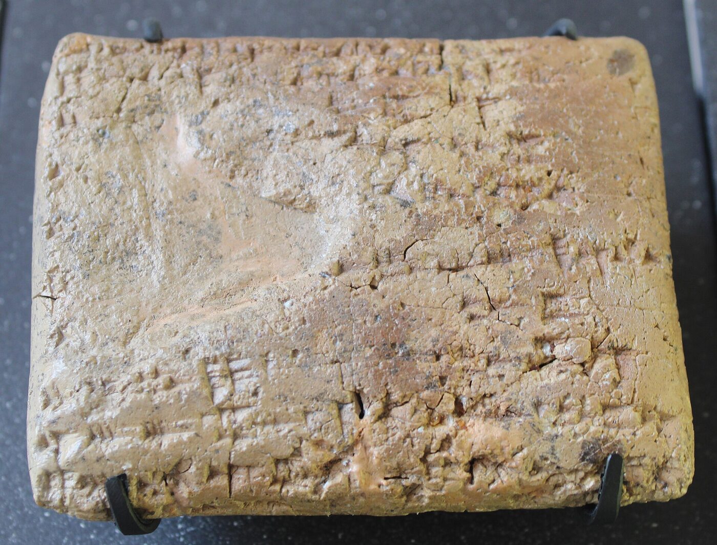 A worn rectangular clay tablet held in a small museum mount and photographed from above, its surface cracked and covered in rows of wedge-shaped alphabetic signs.
