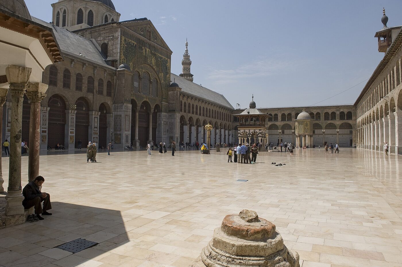 A wide paved courtyard enclosed by two-tier arcades, with the mosaic-faced prayer-hall front and a minaret at the left, a domed treasury raised on columns and a smaller domed kiosk at the right, and visitors crossing the pale stone floor.