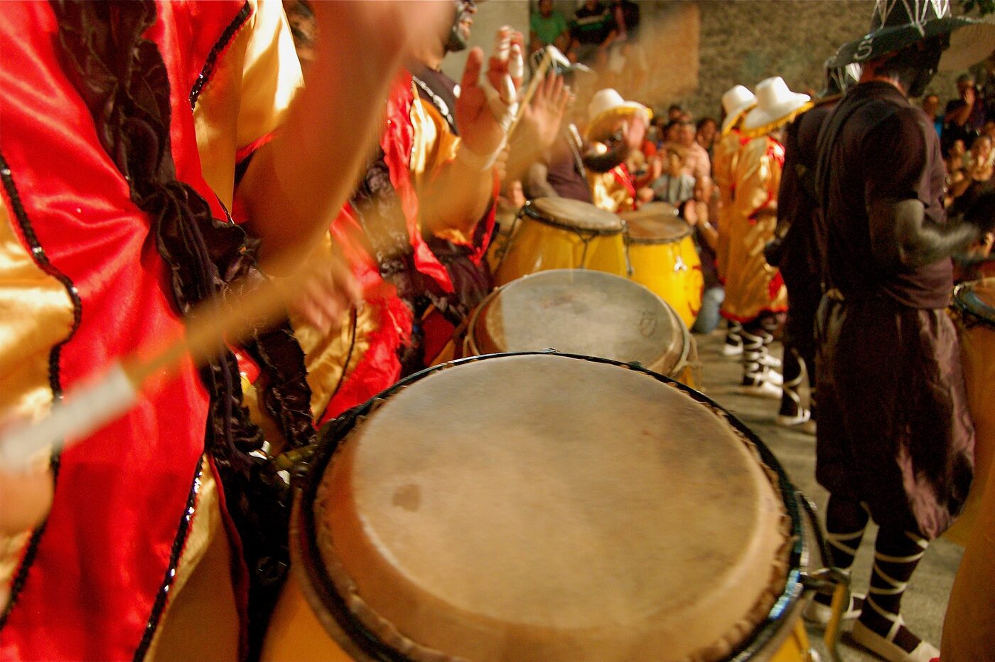 Close view along a line of barrel-shaped drums carried at waist height in a night street parade, hands and sticks blurred in mid-stroke, drummers in red and gold costume at the left.