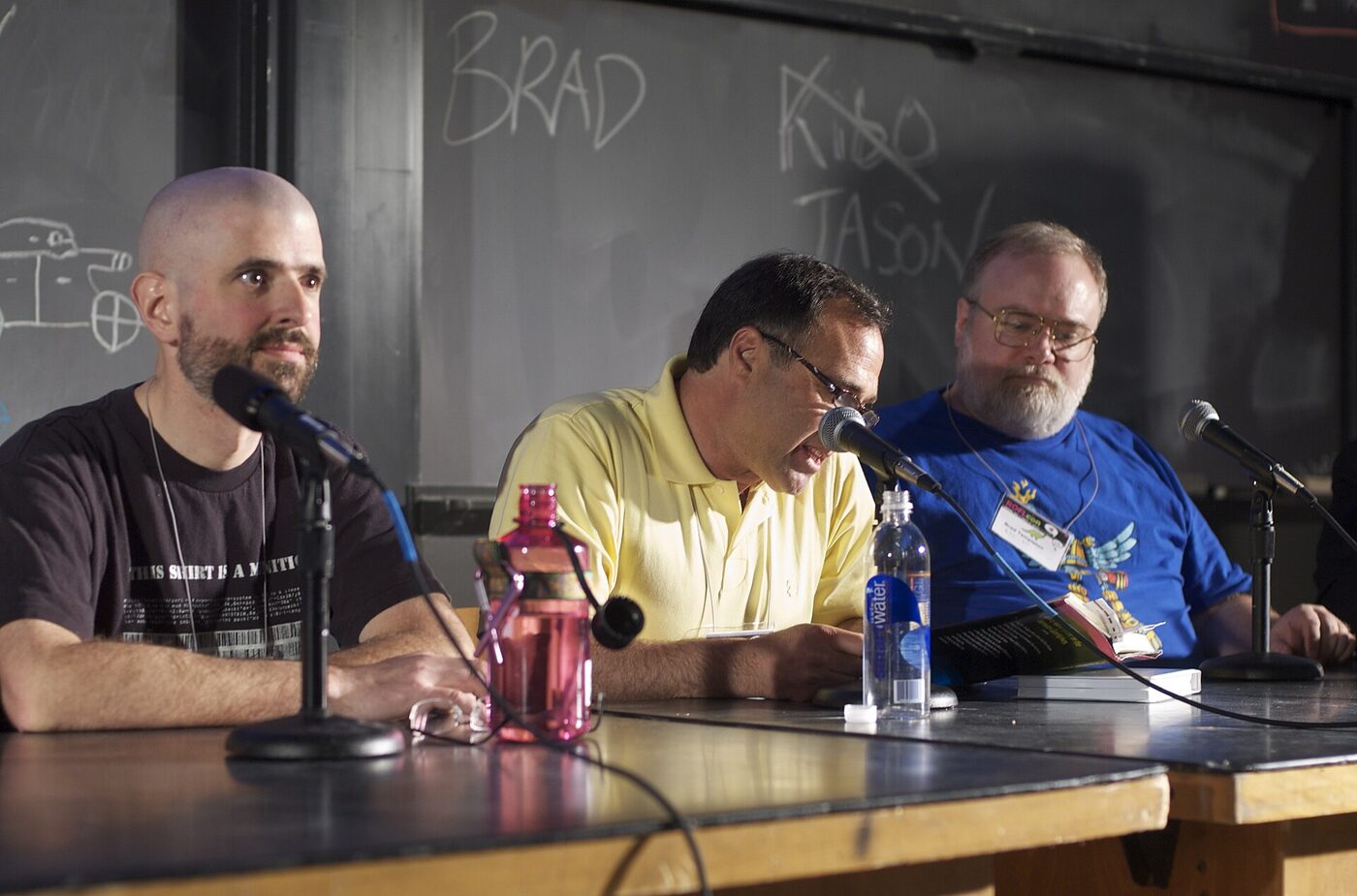 Three men seated behind microphones at a long table in front of a blackboard, one speaking into a microphone while the others listen.