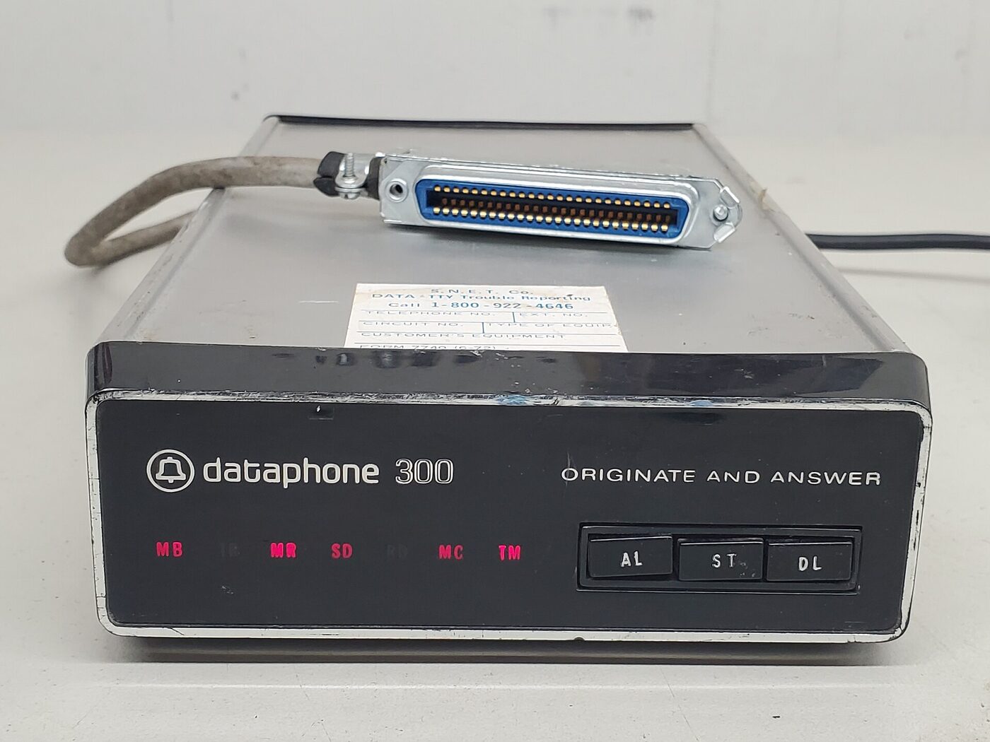 A black-and-chrome desktop modem lying on a pale surface, its front panel lettered “dataphone 300” beside a Bell System logo and the words “originate and answer”, with a row of small red indicator lamps and three push-buttons marked AL, ST and DL; a thick multi-pin connector and cable rest on top of the case.